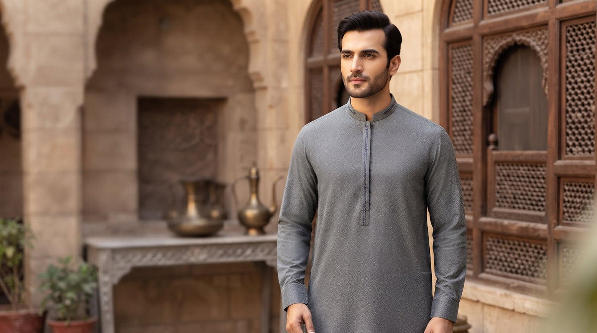 A man wearing a gray long-sleeve kurta stands in front of a traditional stone courtyard with wooden lattice windows and arched alcoves.